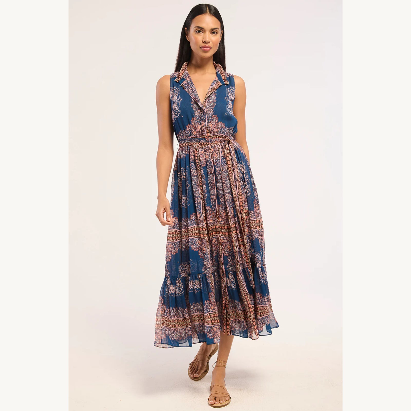 A woman modeling a sleeveless midi dress with a blue base and a detailed brown and beige bohemian pattern, set against a plain light background.