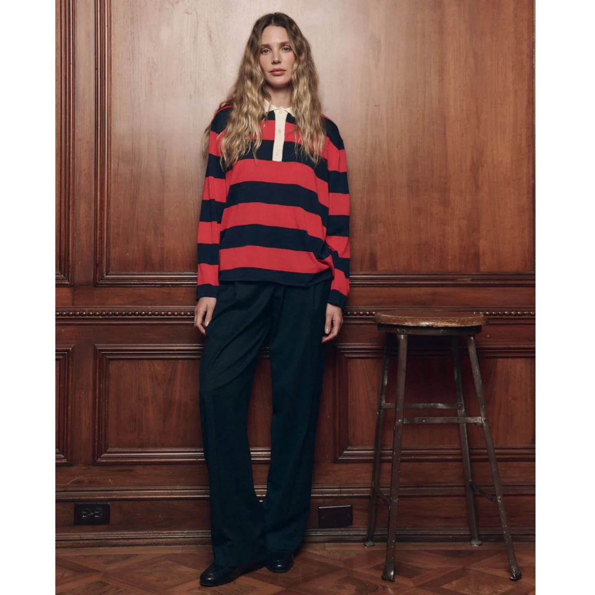 A woman stands in a room with dark wood paneling and a wooden stool, wearing a red and navy horizontally striped long-sleeve rugby-style sweater with a white placket, paired with dark wide-leg trousers.