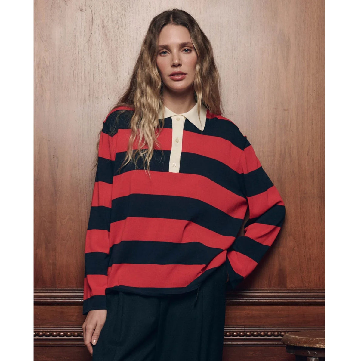 A woman wearing a red and navy blue horizontally striped long-sleeve rugby shirt with a cream collar and placket, paired with dark trousers, standing against a warm wood-paneled wall.