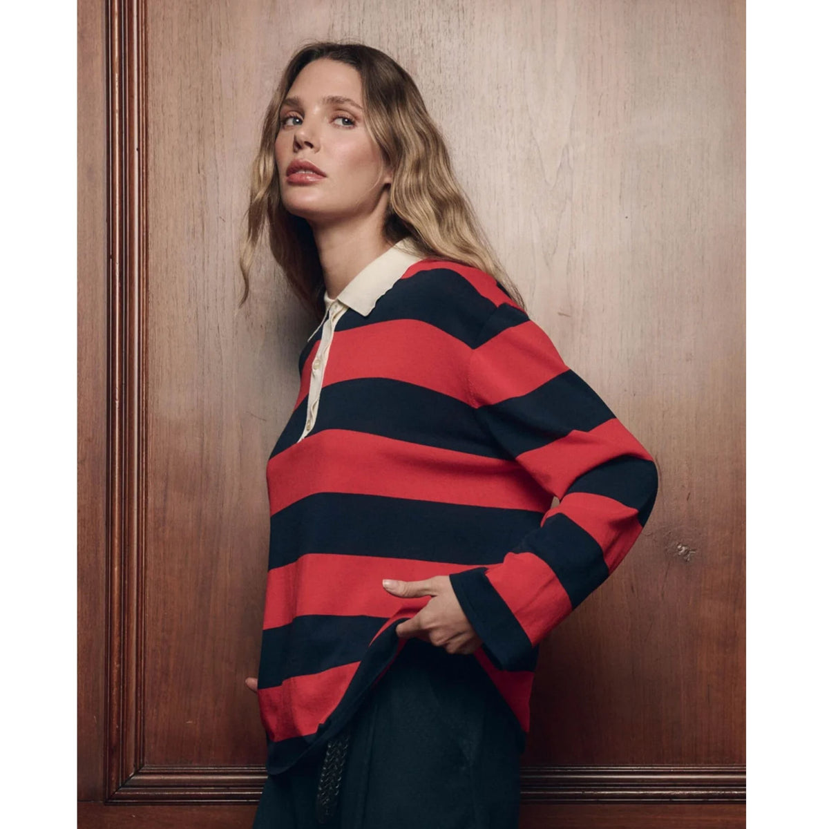 A woman wearing a red and navy blue horizontally striped long-sleeve rugby shirt with a cream collar, leaning against a dark wood-paneled wall.