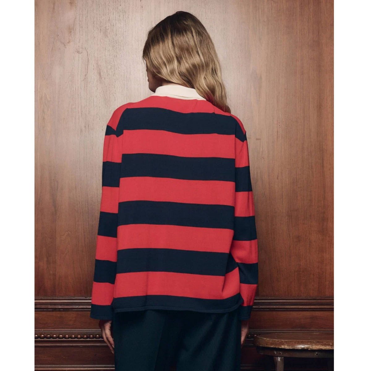 A woman viewed from behind wearing a red and navy blue horizontally striped knit sweater, standing in front of a wooden wall.