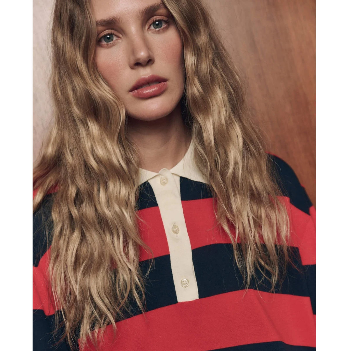 A close-up portrait of a woman with long wavy blonde hair wearing a striped knit top with horizontal red, navy, and cream stripes, set against a warm brown background.