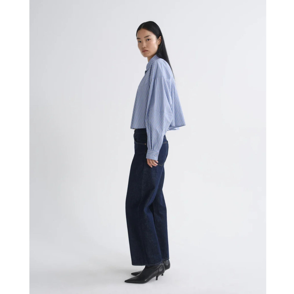 Full-length side profile view of a model wearing the blue and white striped AGOLDE Agnes Shirt and dark navy pants against a light gray background.
