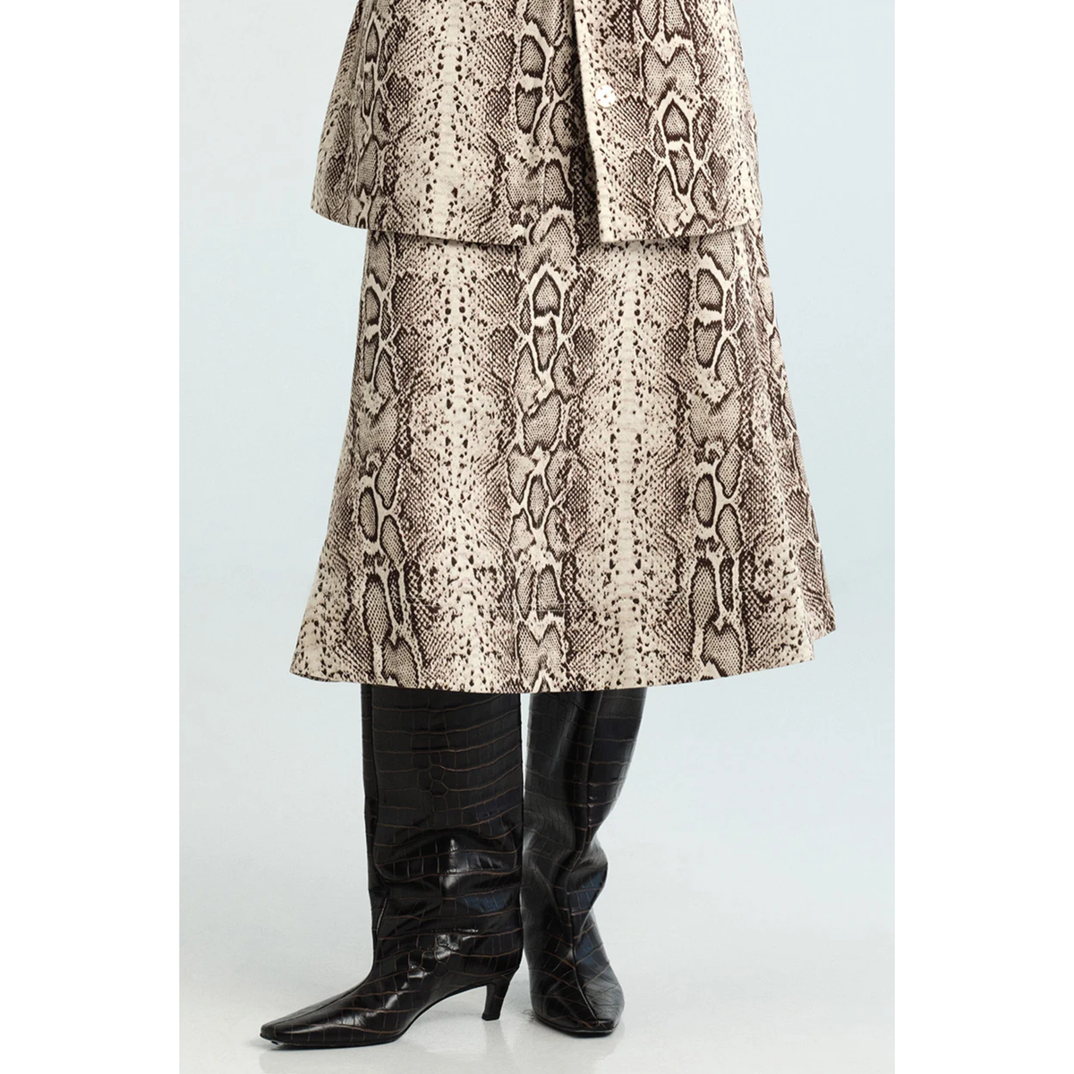 A person wearing a beige and brown snakeskin-patterned skirt or duster with black pointed-toe heeled boots, shown from the waist down against a light gray studio background.