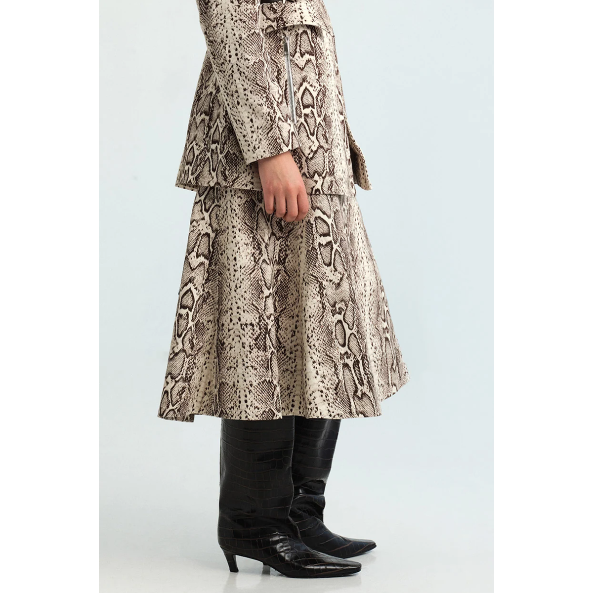 A person wearing a beige and brown snakeskin print skirt with a matching jacket, paired with black knee-high boots, shown against a light gray studio background.