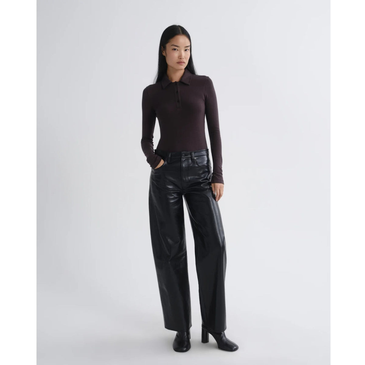 Full-length view of a model wearing a dark brown long-sleeve polo shirt, black leather trousers, and dark boots against a light gray background.
