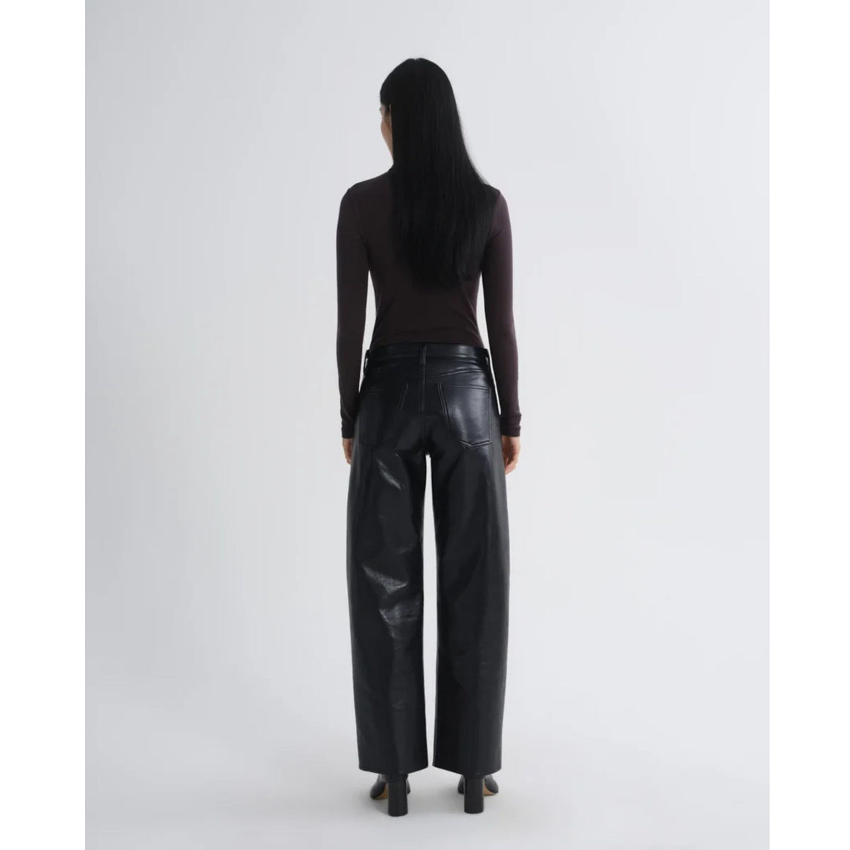 Back view of a model with long dark hair wearing a fitted dark brown long-sleeve top and black leather pants against a light gray background.
