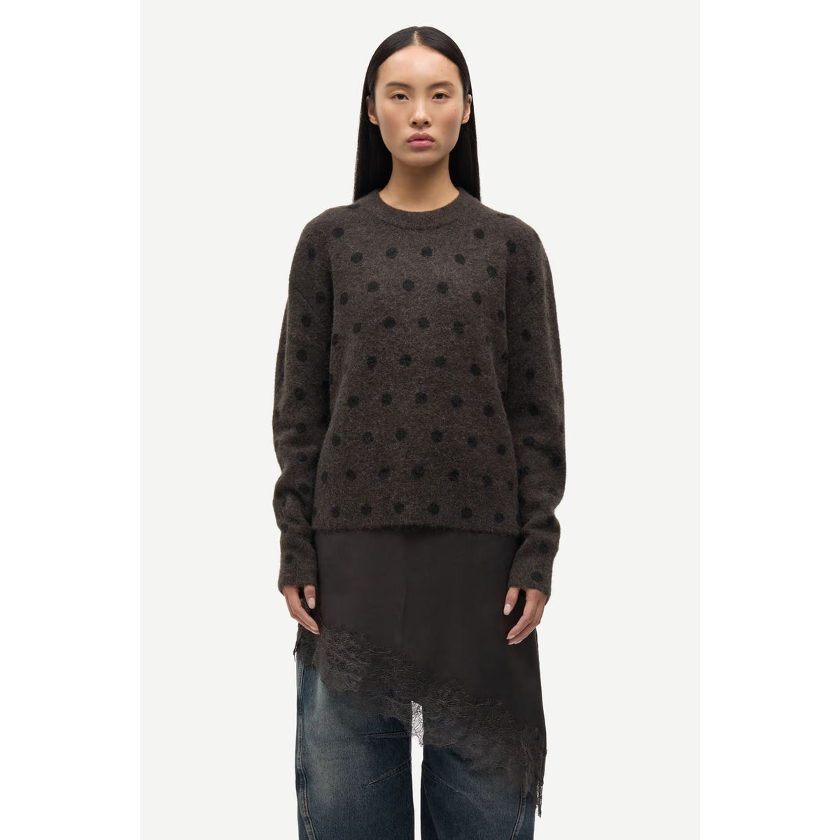 A model wearing a dark brown crewneck sweater with a subtle black polka dot pattern, paired with a black asymmetrical draped tunic and blue jeans, set against a plain light gray background.
