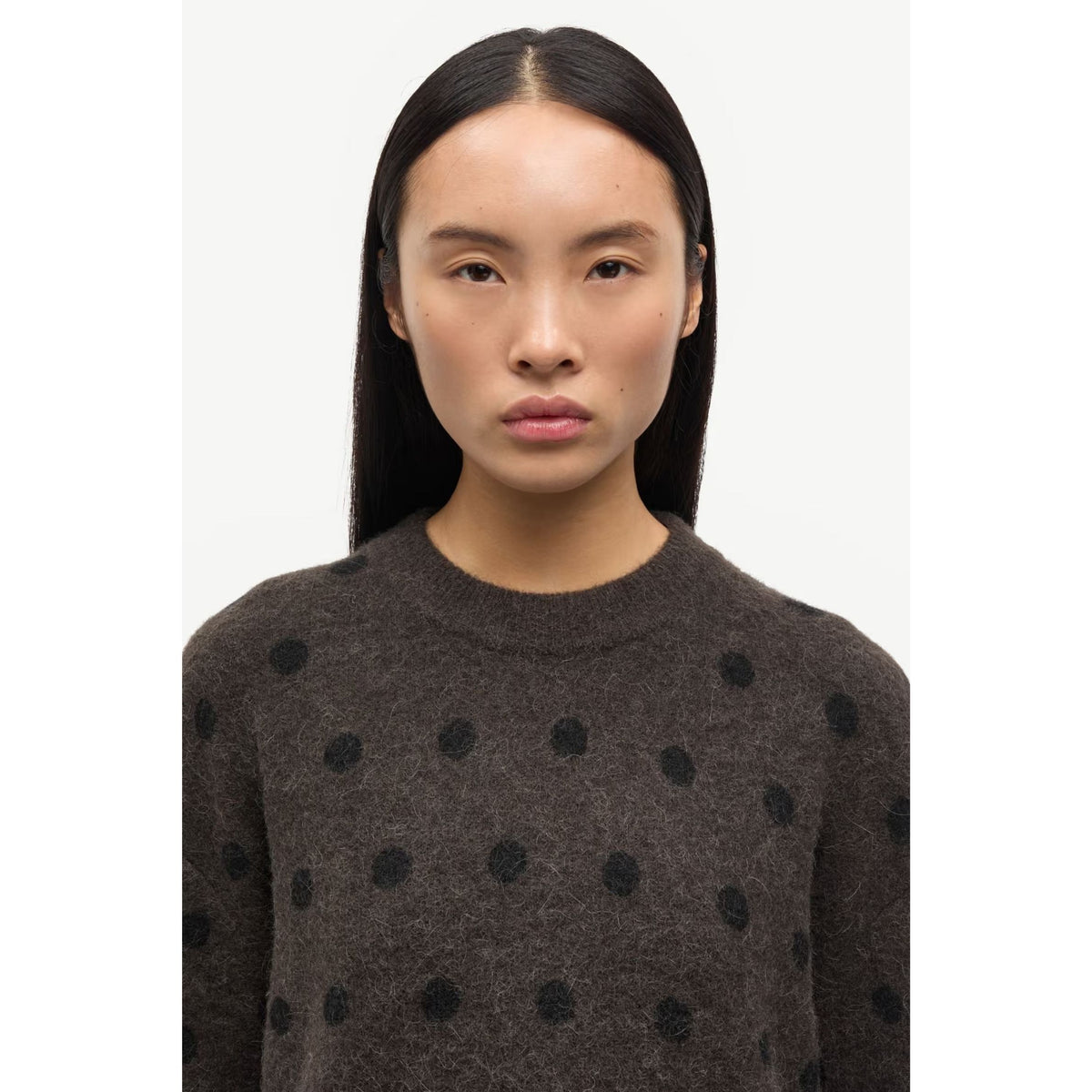 A woman with long, straight black hair wearing a dark brown crewneck sweater with black polka dots, photographed against a plain light gray background.