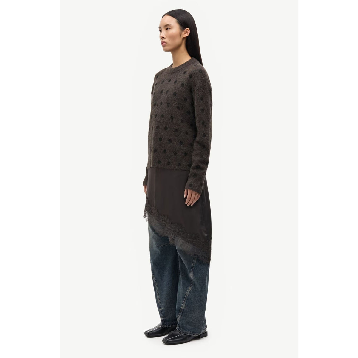 A model is shown in profile wearing a dark brown textured knit tunic sweater with a sheer overlay and wide-leg blue jeans against a plain light gray background.