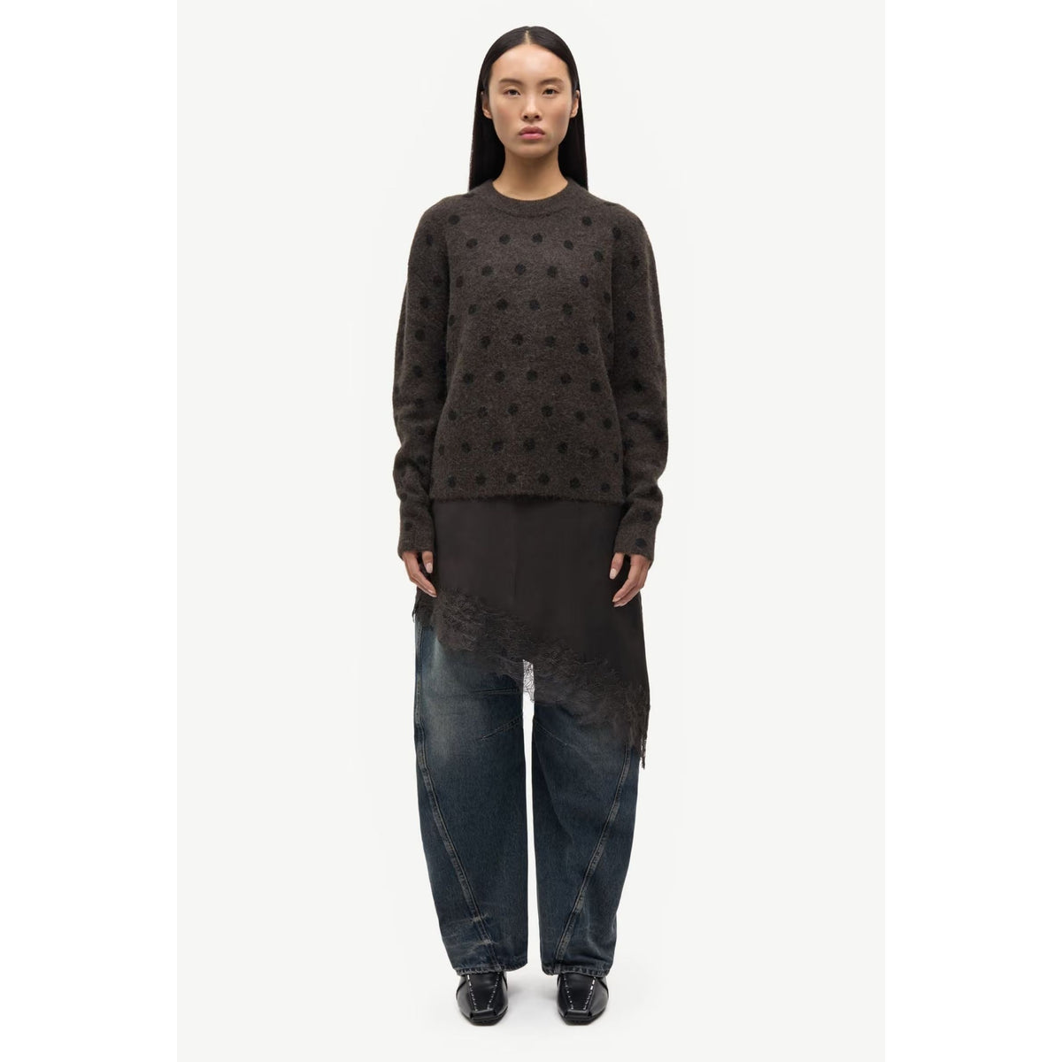 A model stands against a light gray studio background wearing a dark brown knit sweater with a textured dotted pattern, a black asymmetrical draped tunic, wide-leg dark blue jeans, and black shoes.