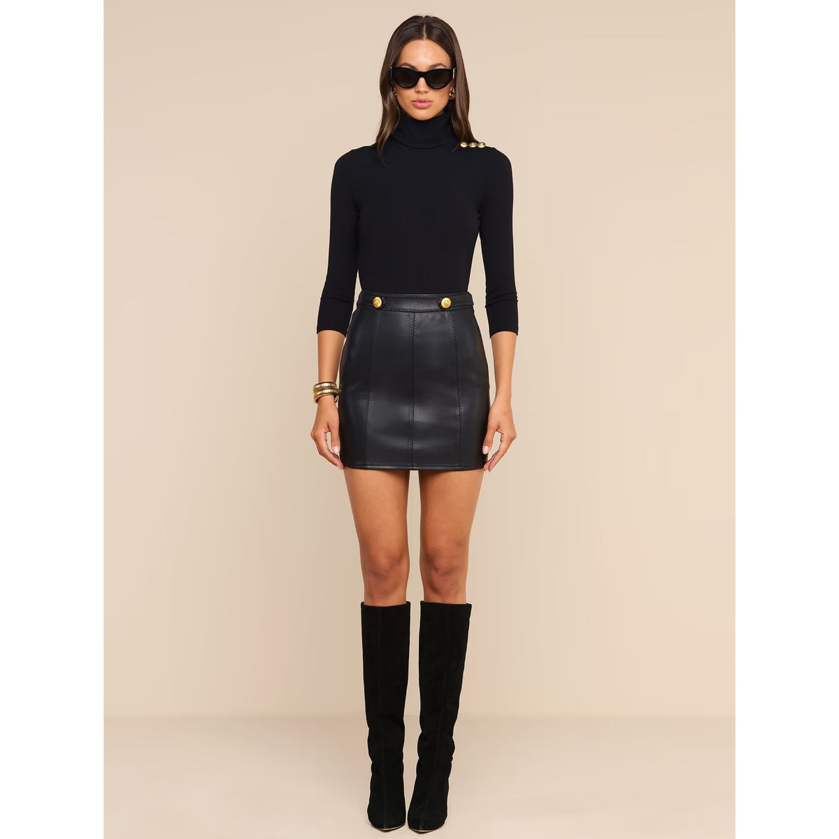 A model wearing a black mock-neck long-sleeve top with gold shoulder buttons, a black faux-leather mini skirt with gold button details at the waist, and black knee-high boots, set against a solid beige studio background.