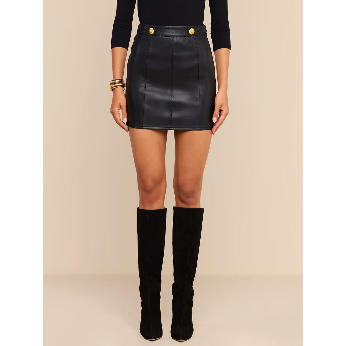 A model wearing a black faux-leather mini skirt with gold button details, paired with a black long-sleeve top and black knee-high boots, set against a solid beige studio background.