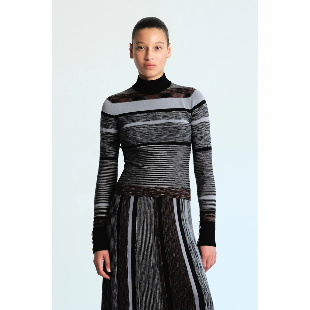 A model wearing a long-sleeve mock neck knit top and matching pleated skirt in black, gray, and white striped textured fabric, set against a light blue studio background.