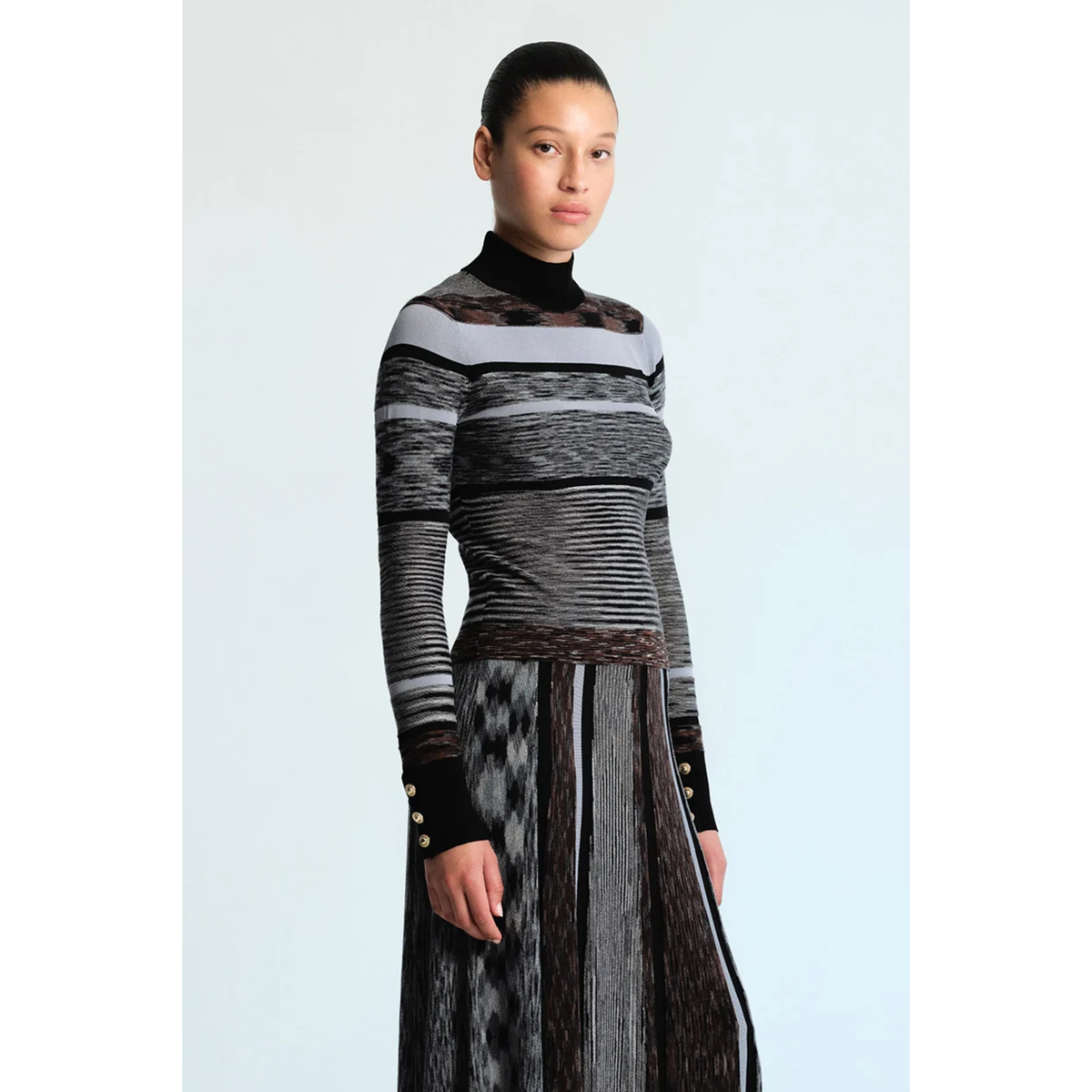 A model wearing a long-sleeve mock-neck knit dress with a black, gray, and white patterned design, set against a light blue studio background.