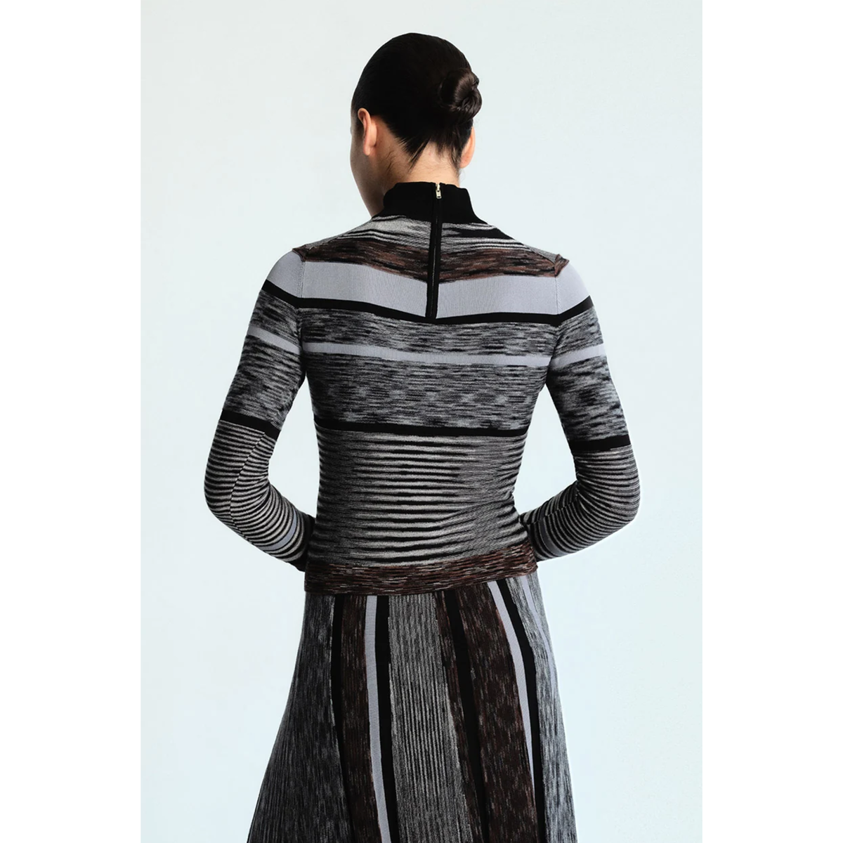 A model viewed from the back wearing a long-sleeve mock-neck knit top and matching pleated skirt with horizontal and vertical striped patterns in black, gray, white, and brown.