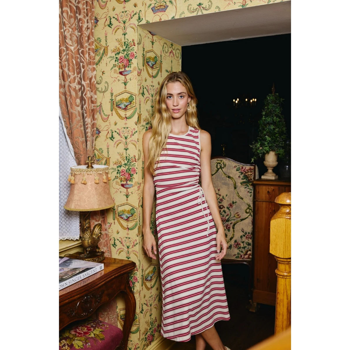 A woman modeling a sleeveless, halter-style midi dress with red and white horizontal stripes, standing in a vintage room with ornate wallpaper.