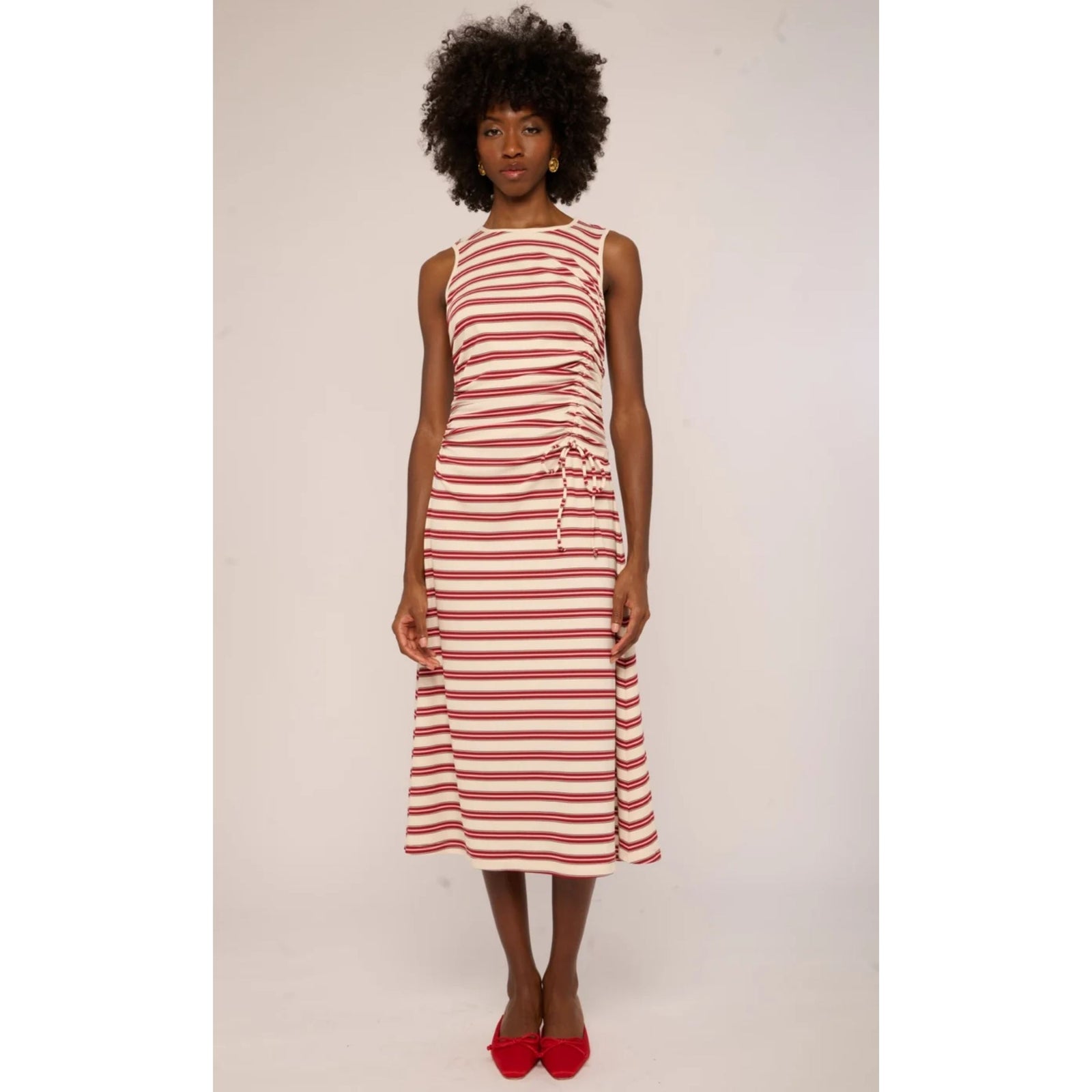 A woman modeling a sleeveless, knee-length dress with horizontal red and off-white stripes, featuring a tie-waist detail and red pointed-toe heels, set against a plain light background.