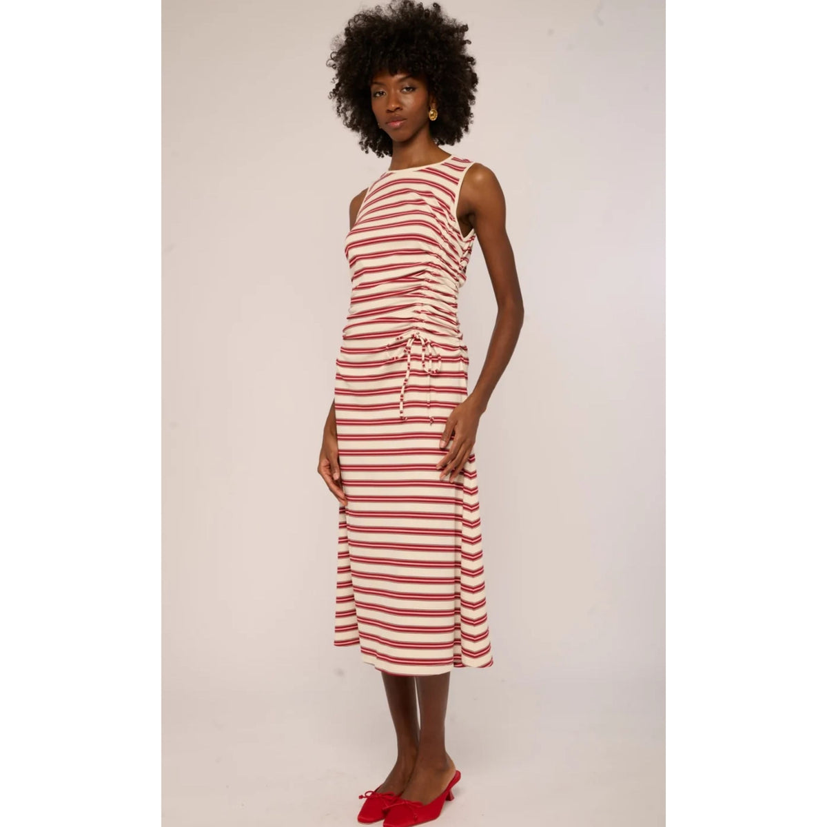 A woman modeling a sleeveless midi dress with horizontal red and off-white stripes, featuring a gathered waist and tie detail, paired with red pointed flats against a plain light background.