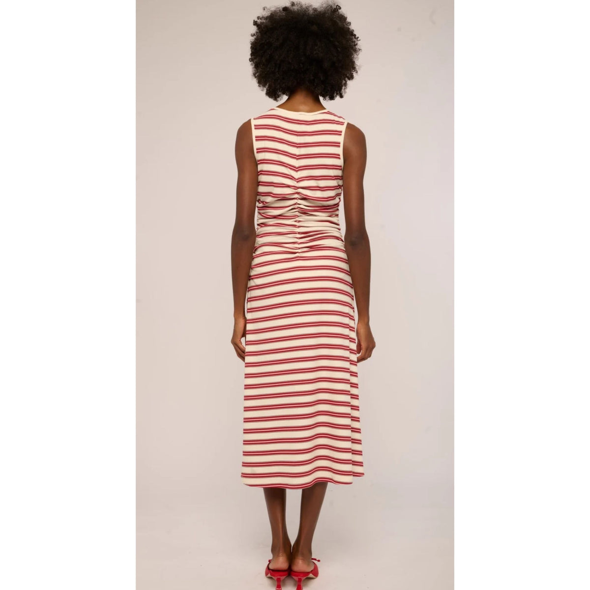 A woman viewed from the back wearing a sleeveless midi dress with horizontal red and off-white stripes, paired with red high heels, against a plain light background.