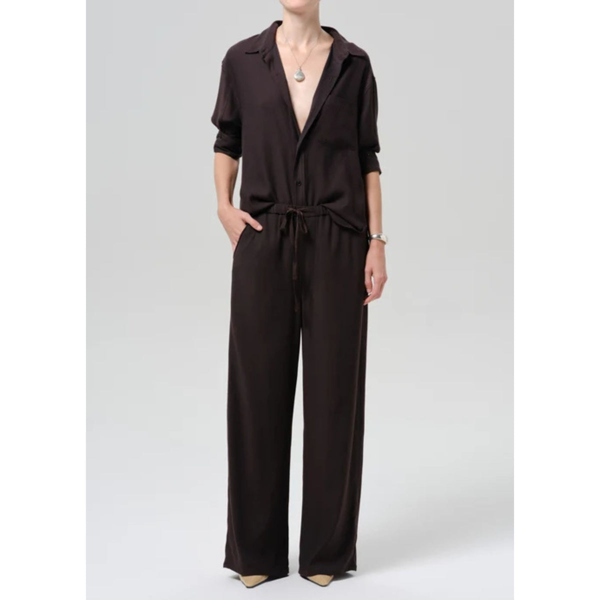 A model wearing a dark brown collared jumpsuit with a drawstring waist and wide legs, photographed against a neutral light gray studio background.