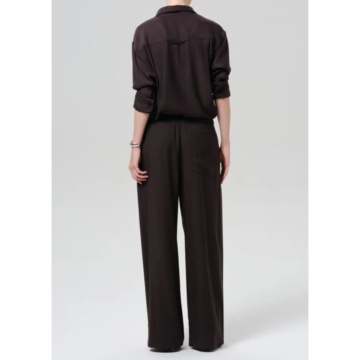 A model shown from the back wearing a dark brown long-sleeve button-up shirt and matching dark brown wide-leg trousers, set against a plain light gray studio background.