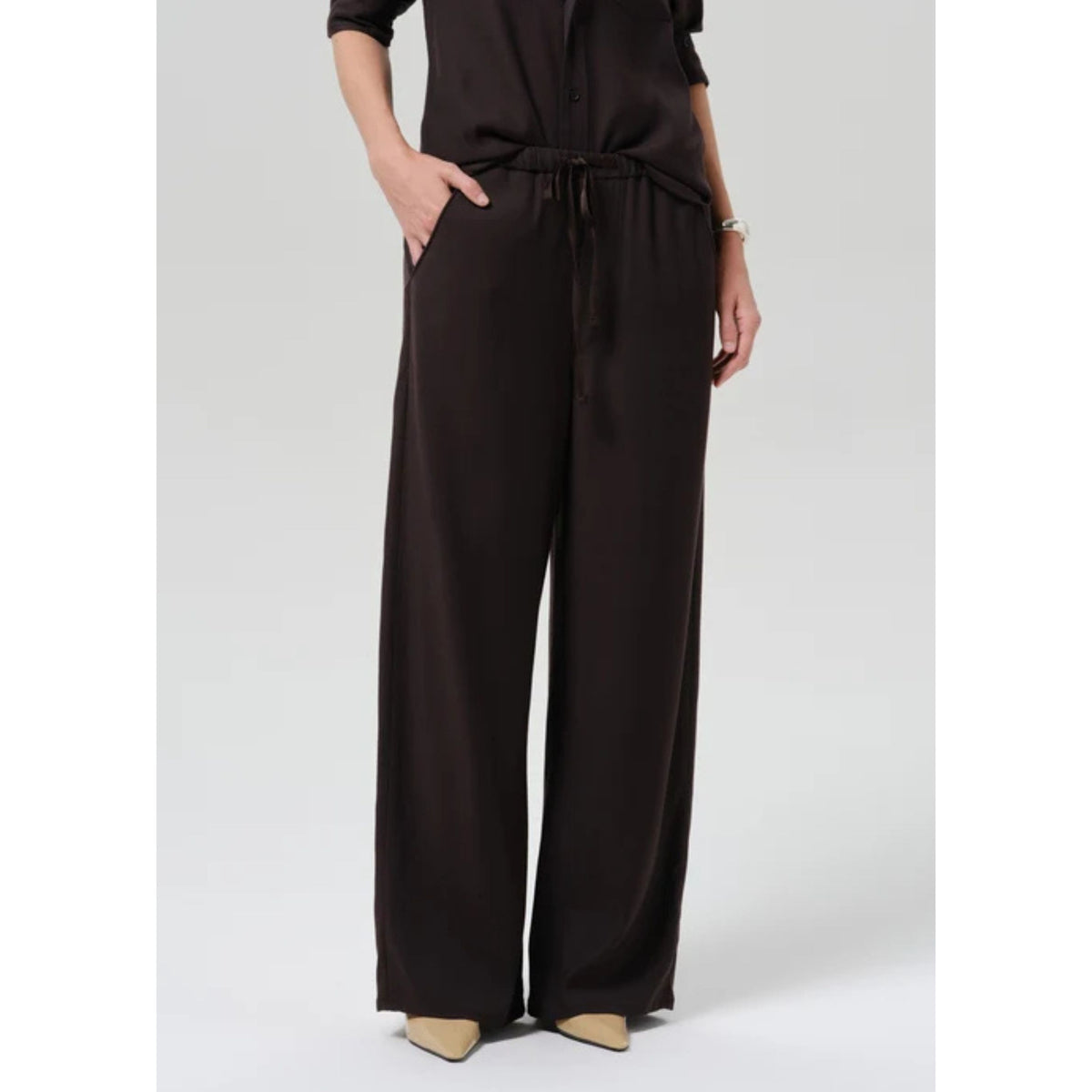 A model wearing dark brown wide-leg trousers with a drawstring waist, paired with a matching dark brown top and beige pointed-toe heels, shown against a light gray studio background.