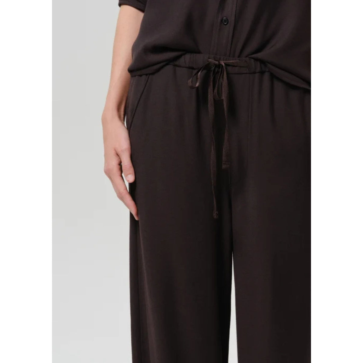 A close-up of a person wearing dark brown wide-leg trousers with a matching drawstring waist and a dark brown top, shown against a light gray studio background.