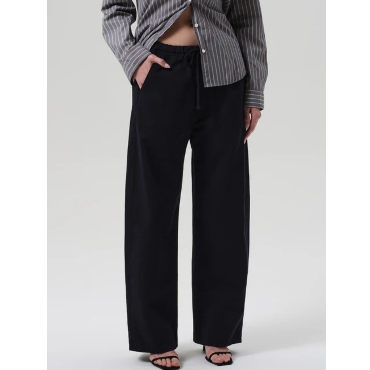 A model wearing black wide-leg drawstring pants, paired with a gray and white striped button-up shirt and black sandals, shown from the waist down against a light gray studio background.
