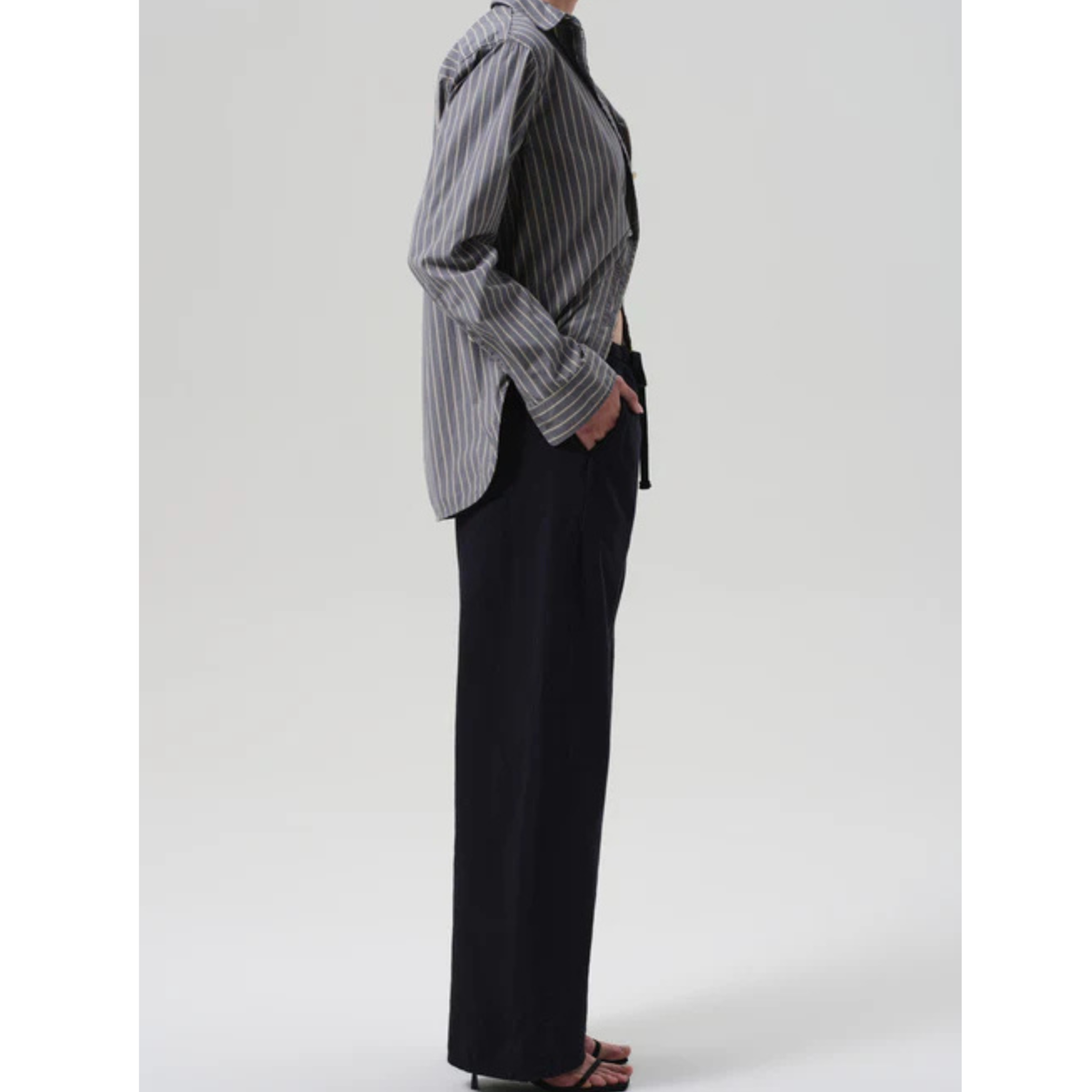 A model shown from the side wearing a black and white vertically striped button-down shirt and black wide-leg trousers, standing against a light gray studio background.