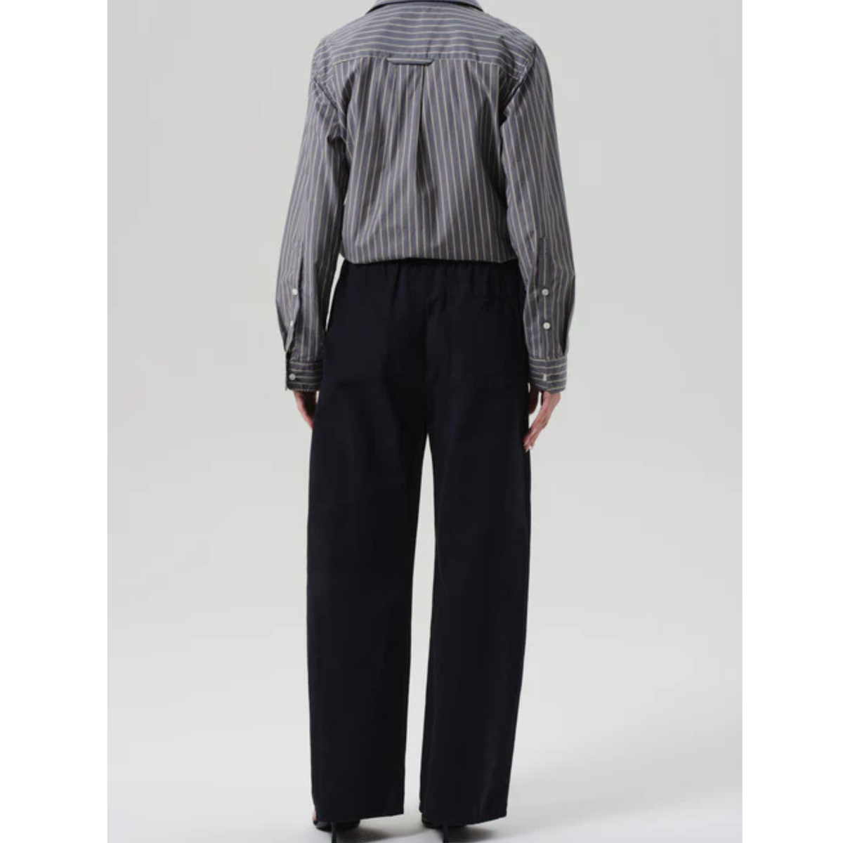 A person shown from the back wearing a gray and black vertically striped button-up shirt and black wide-leg trousers, photographed against a light gray studio background.