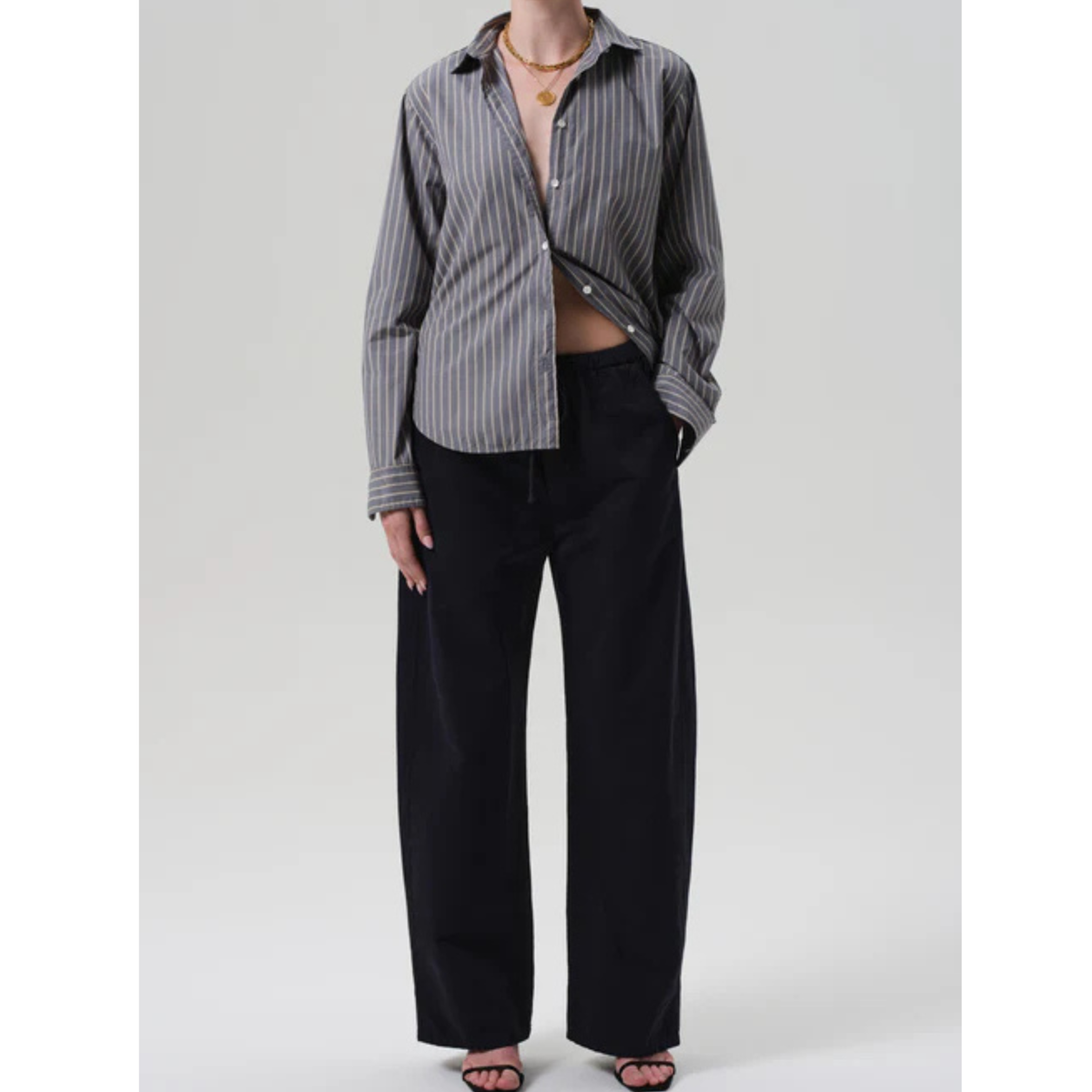 A model wearing a gray and black vertically striped button-down shirt, styled open over a bare midriff, paired with black wide-leg pants and black flat sandals, against a light gray studio background.