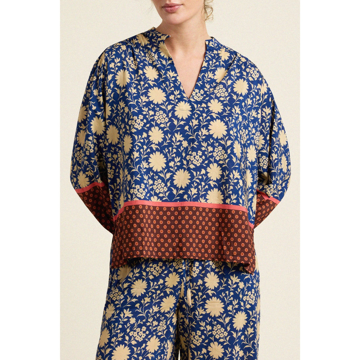 A woman wearing a blue and beige floral print top with a brown patterned waistband and pink trim, paired with matching floral pants, against a light beige background.
