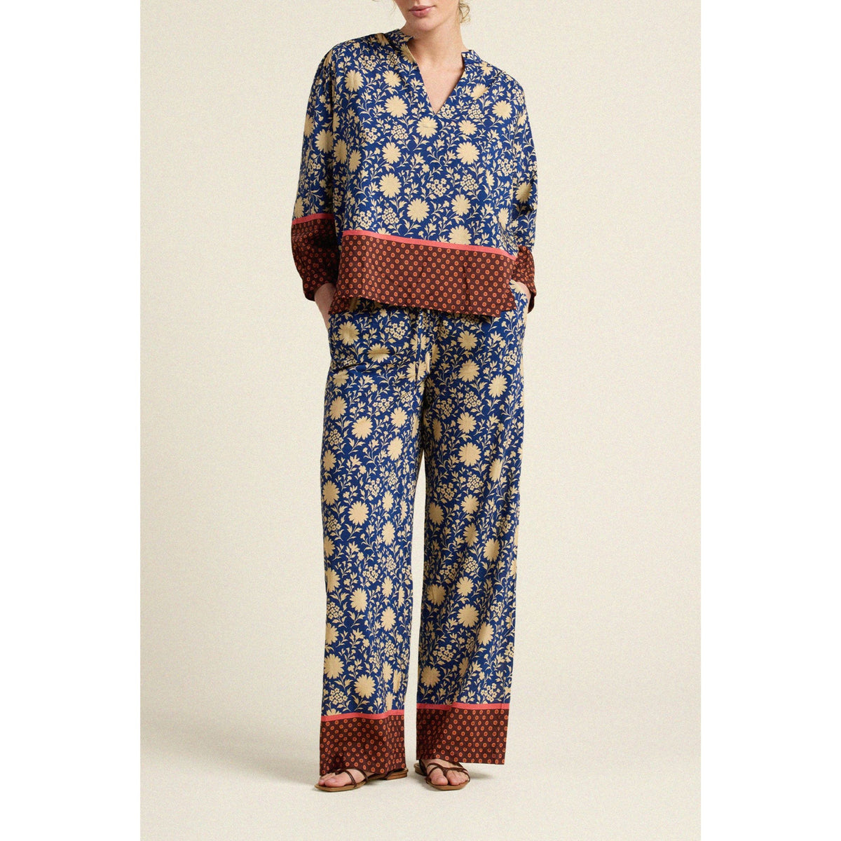 A woman modeling a matching two-piece pajama set with a blue and beige floral print and contrasting red patterned cuffs and hem, photographed against a plain beige studio background.