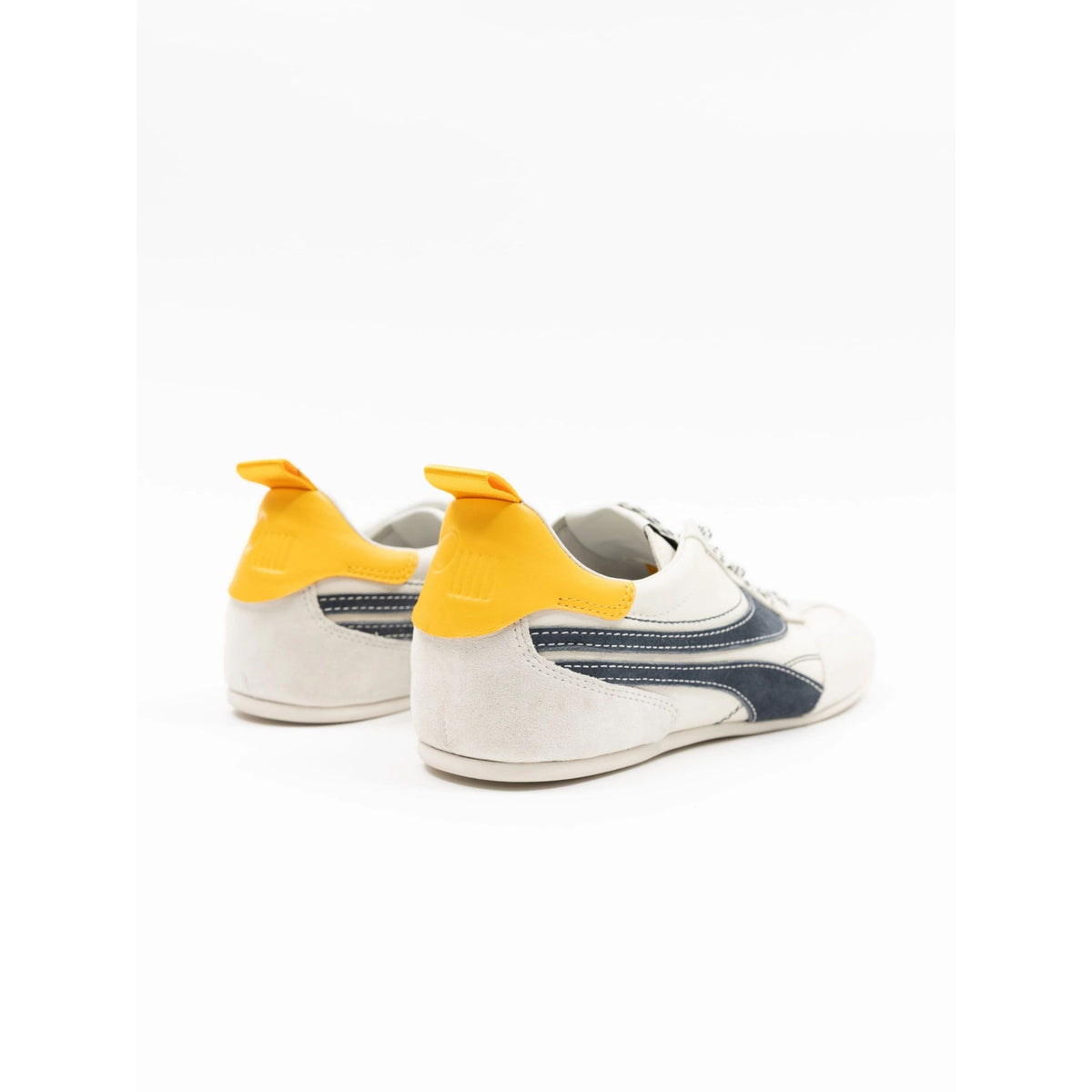 A pair of low-top sneakers shown from the rear three-quarter angle on a clean white background. The shoes feature a cream suede upper, a bright yellow heel tab, and a navy blue side stripe.