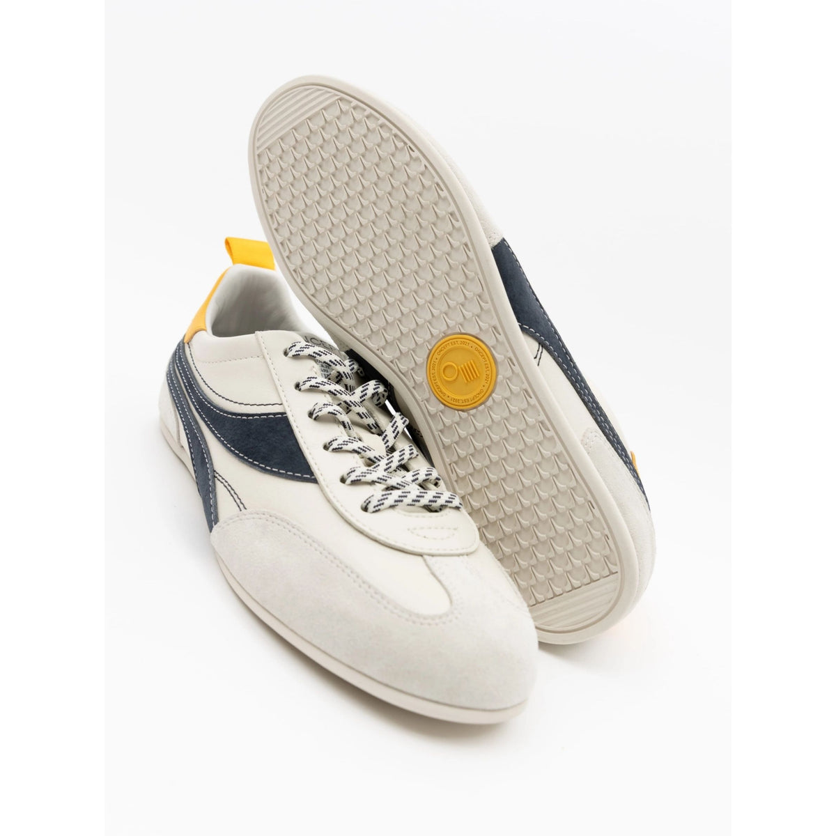 A pair of low-top sneakers shown on a clean white background, with one shoe standing upright to display the textured outsole and the other lying on its side showing the side profile. The shoes are primarily off-white suede and leather with navy blue side stripes, white-and-black patterned laces, and a small yellow accent on the heel tab and circular outsole logo.