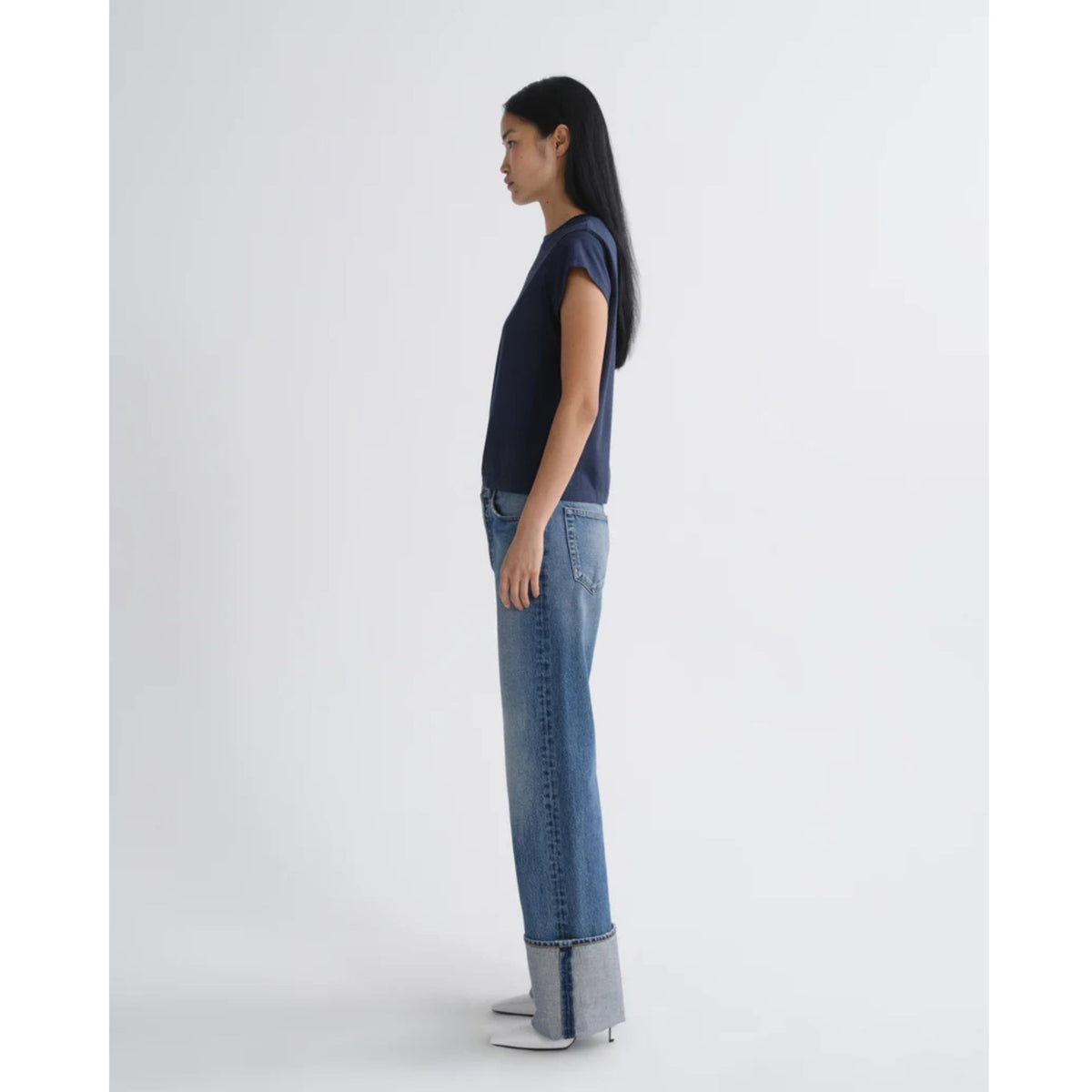 Side profile view of a model wearing a navy short-sleeve top and AGOLDE Big Cuff Jeans in Liberty with large cuffed hems against a light gray background.