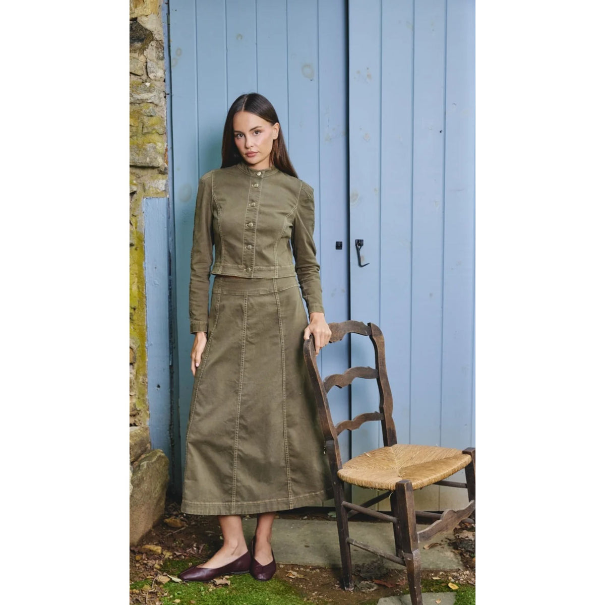 A woman models an olive green two-piece set consisting of a long-sleeve button-front top and matching midi skirt, standing next to a wooden chair in front of a blue wooden door.