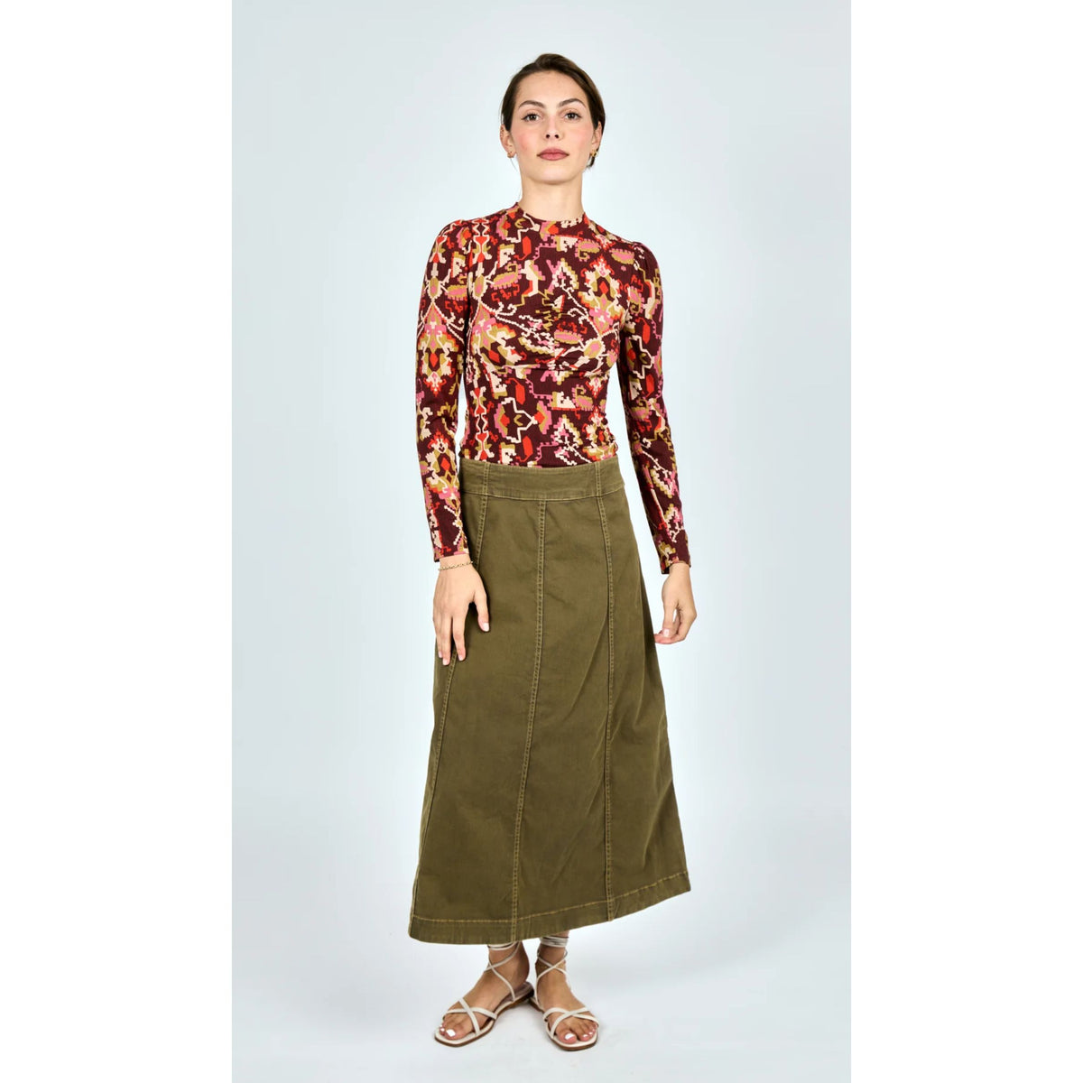 A woman modeling a long-sleeve mock-neck top with a dense abstract floral pattern in red, orange, yellow, and green, paired with an olive green wrap-style midi skirt and strappy flat sandals, set against a plain light gray studio background.