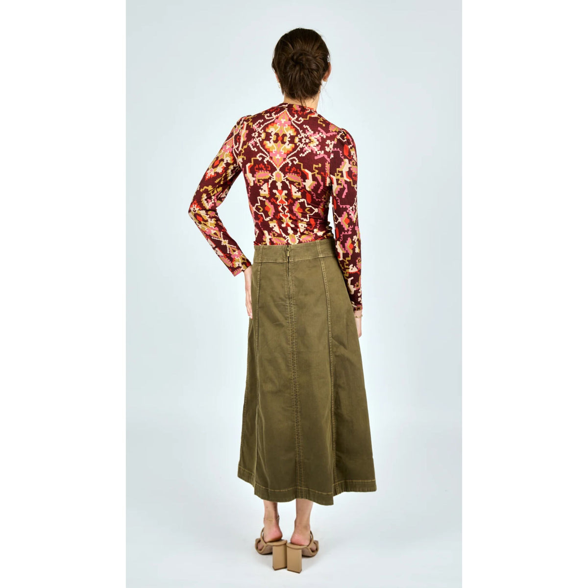 A model is shown from the back wearing a long-sleeve high-neck blouse with a red, orange, and yellow floral/tribal pattern, paired with an olive green midi skirt and brown block-heel sandals, set against a light gray studio background.
