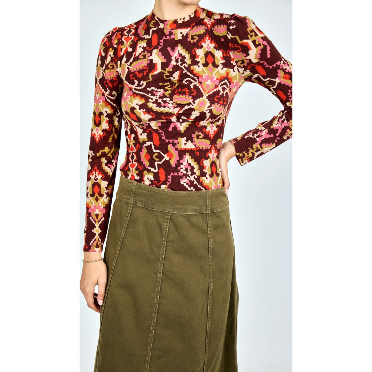 A woman wearing a long-sleeve mock-neck top with a complex multicolor geometric and tribal pattern in red, pink, orange, and yellow, paired with an olive green skirt.