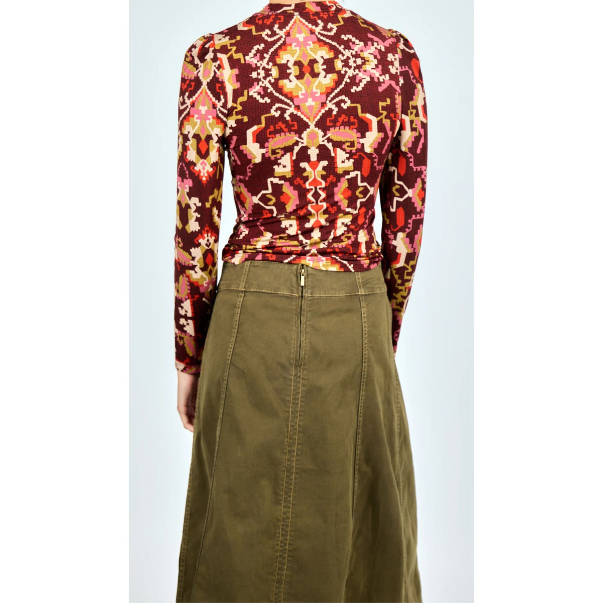 A model shown from the back wearing a long-sleeve fitted top with a red, orange, and yellow geometric pattern, paired with an olive green skirt.
