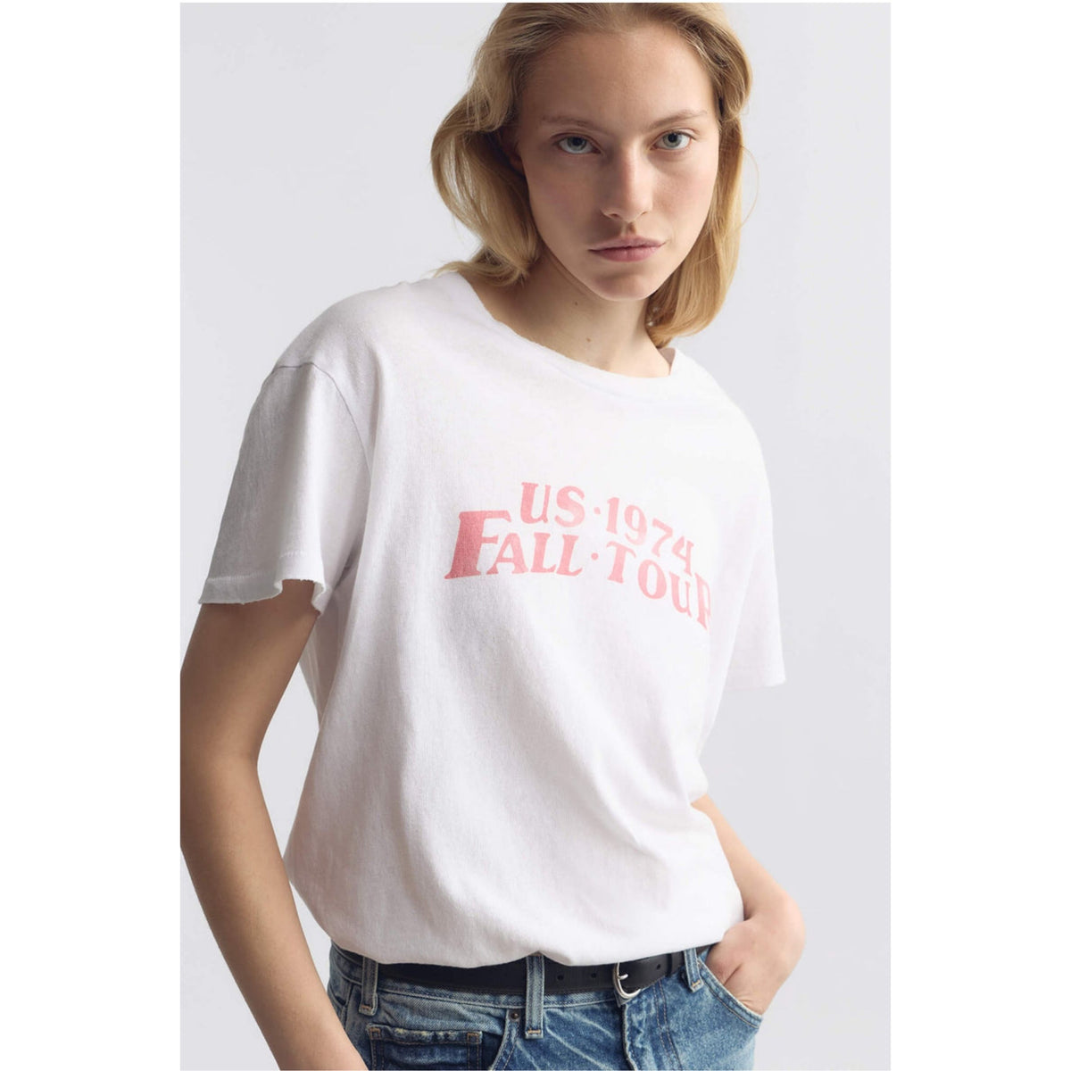 A woman wearing a white short-sleeve t-shirt with a pink graphic text print that reads 'US 1974 FALL TOUR', paired with blue jeans, against a light gray studio background.