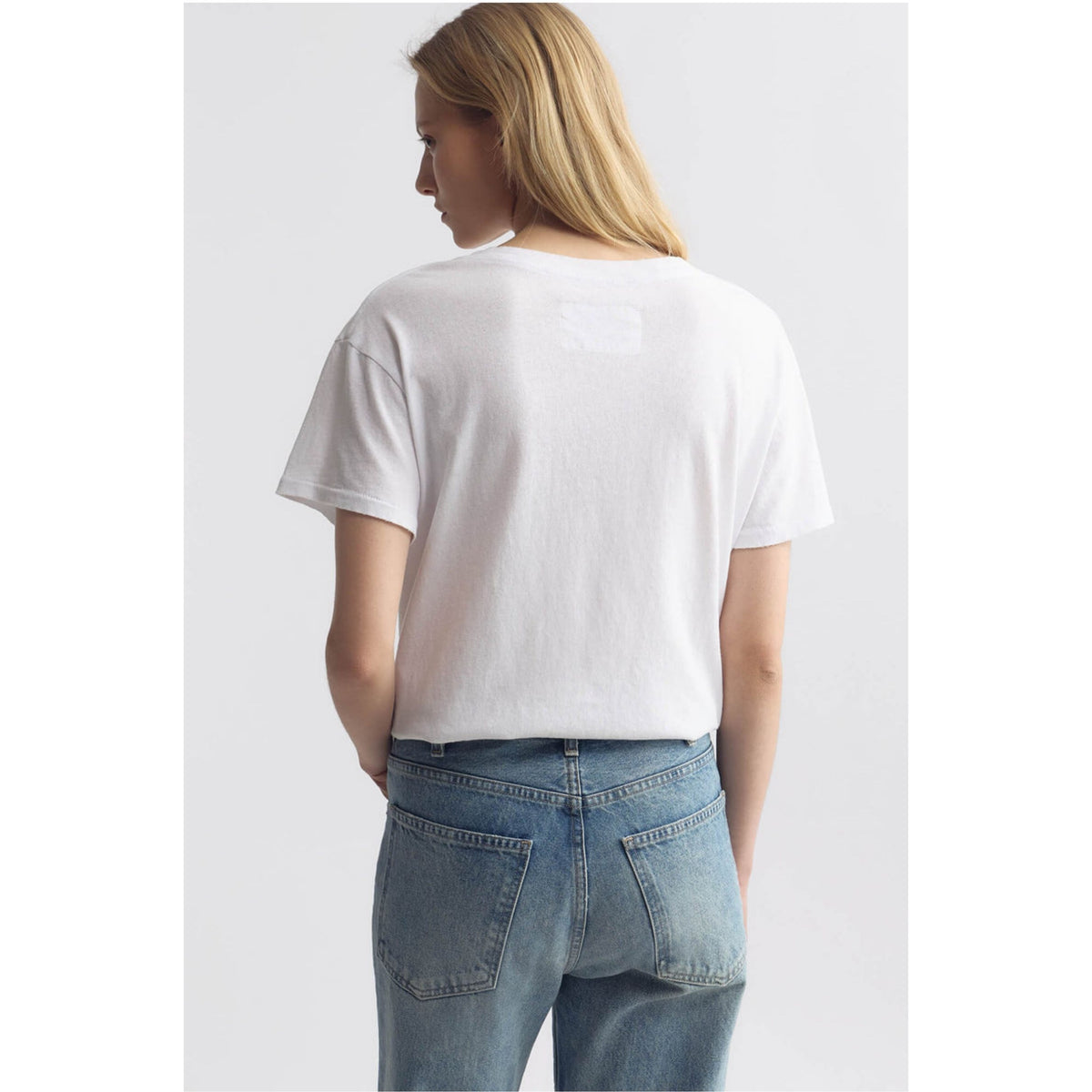 A woman shown from the back wearing a white short-sleeve cropped T-shirt and blue jeans against a light gray studio background.