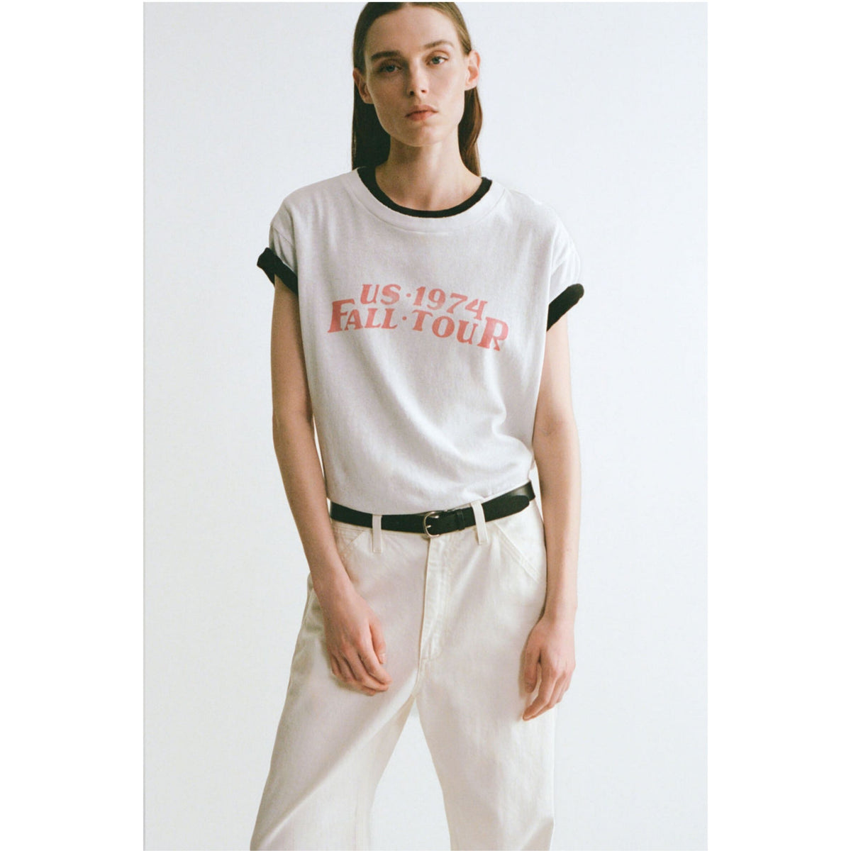 A model wearing a white ringer t-shirt with black ribbed trim and a red graphic print reading 'US 1974 FALL TOUR', paired with white pants and a black belt, against a plain light gray background.
