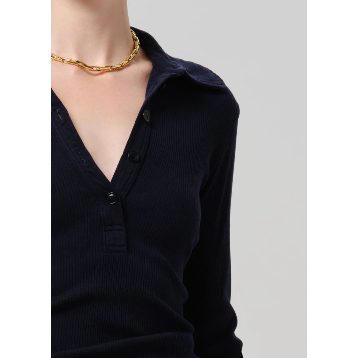 A close-up of a person wearing a dark navy ribbed long-sleeve polo shirt with a button placket and collar, paired with a chunky gold chain necklace, against a light gray background.