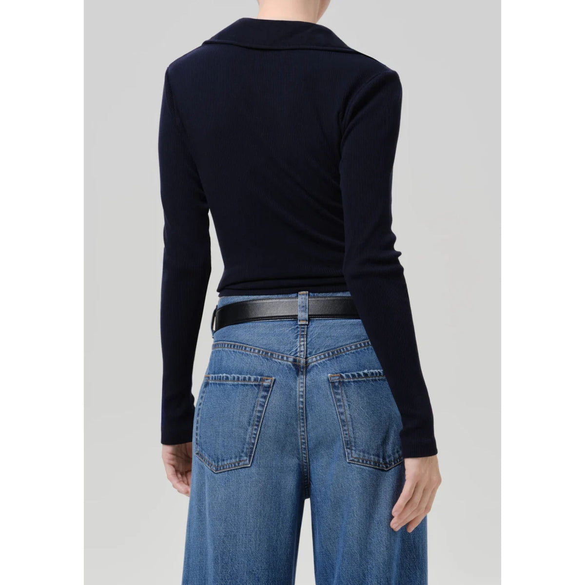 A model shown from the back wearing a dark navy long-sleeve knit top tucked into blue jeans with a black belt, against a light gray studio background.