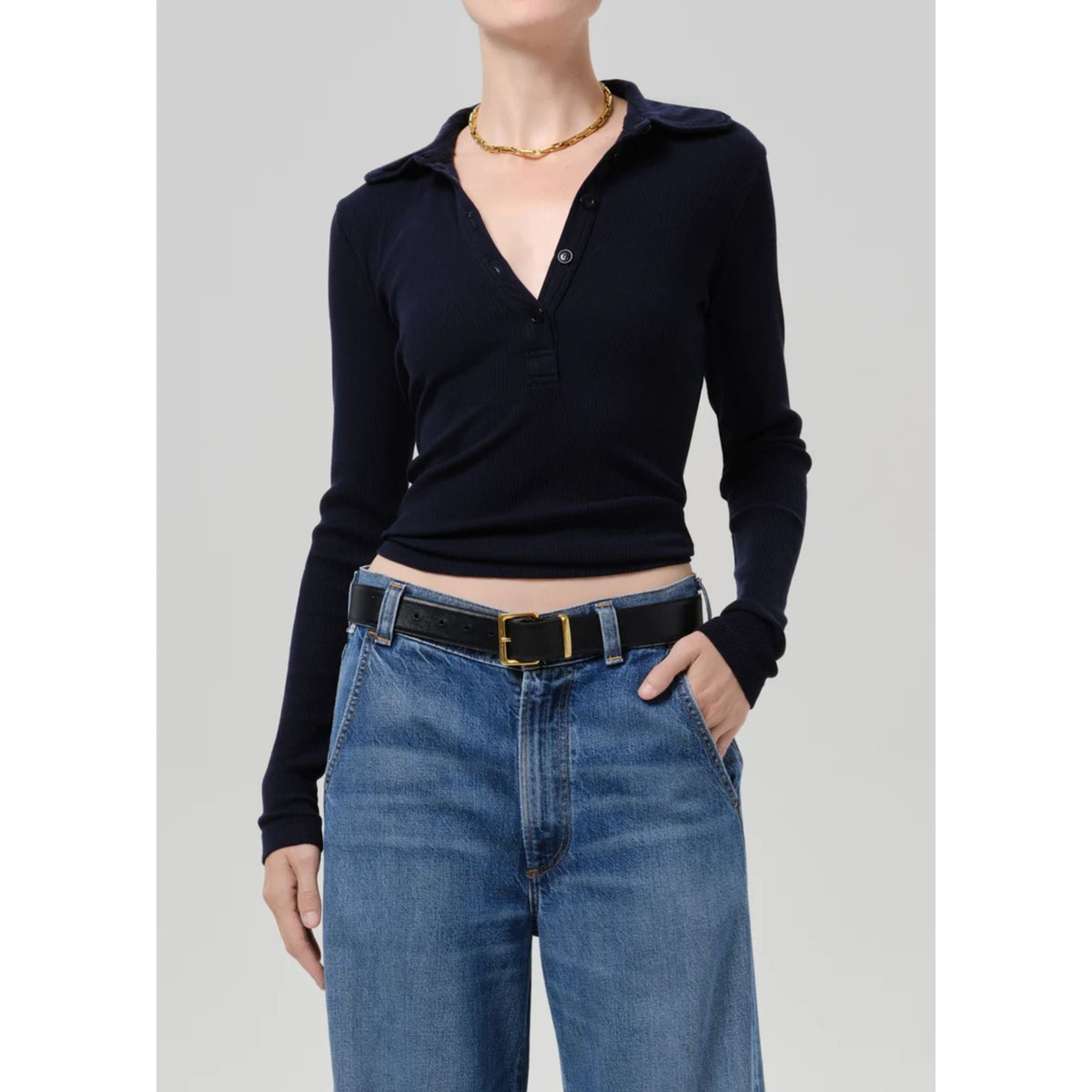 A model wearing a dark navy long-sleeve polo shirt tucked into blue jeans with a black belt, shown from the neck down against a light gray studio background.