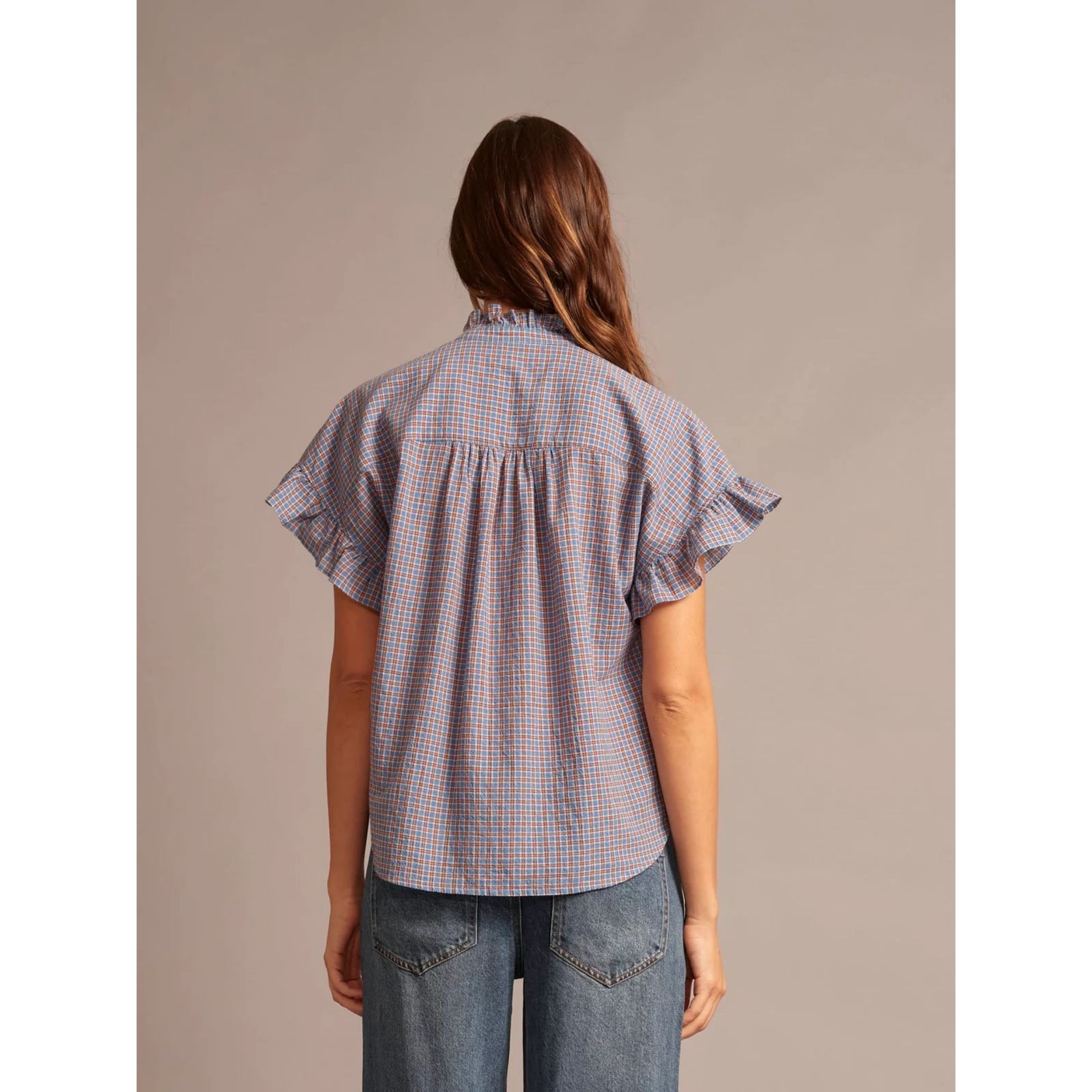 A short-sleeve blouse with a blue, red, and white gingham check pattern, featuring ruffled details on the collar and sleeve cuffs, laid flat against a light gray background.