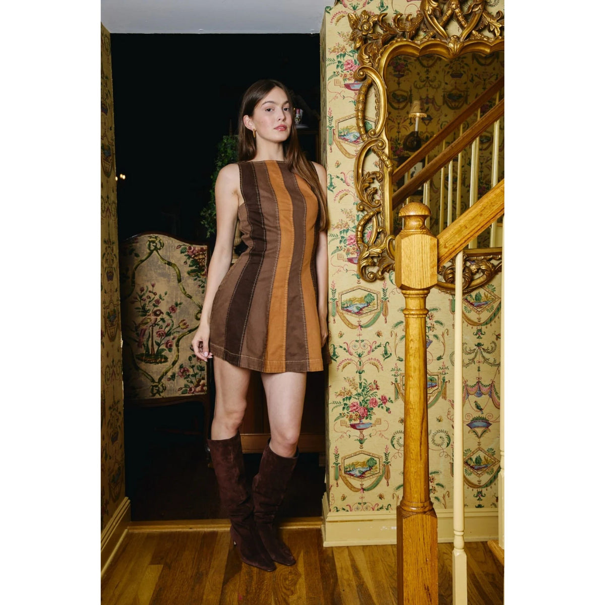 A woman stands in an ornate indoor room wearing a sleeveless brown and tan striped mini dress with matching brown knee-high boots. The background features vintage floral wallpaper, a wooden staircase with gold detailing, and a dark doorway.